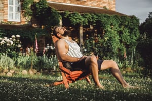 Mann in Sommerkleidung sitzt auf einem Liegestuhl auf einer grünen Wiese vor einem Haus